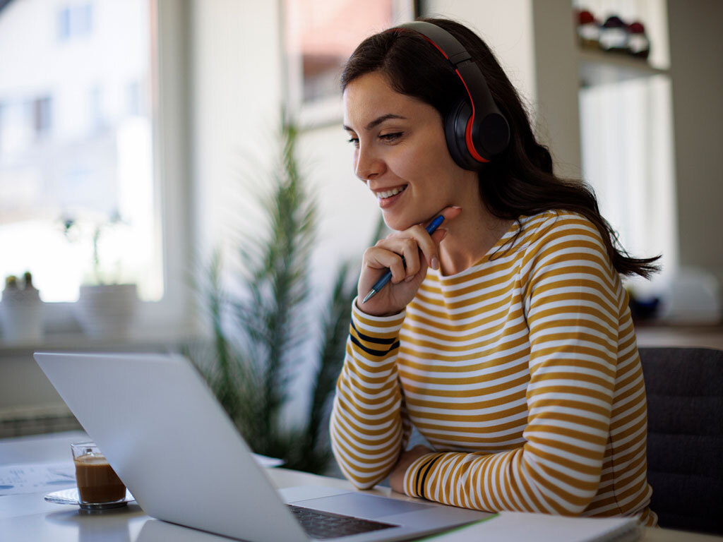 Junge Frau bei der Arbeit im Büro mit Kopfhörern vor einem Laptop, © iStock damircudic